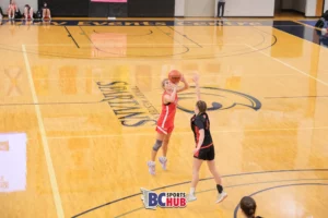 A Flight Basketball athlete is at the start of her jump for a 3-point shot with an incoming defender with her arm up to contest.